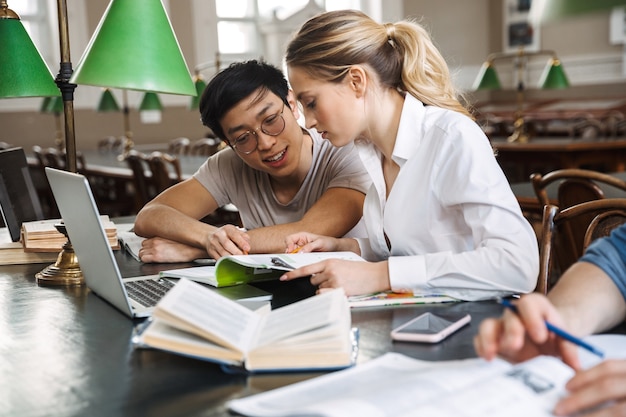 Foto feliz pareja de estudiantes que estudian en la biblioteca mientras está sentado en el escritorio con un portátil