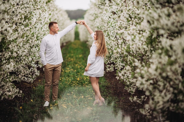 Feliz pareja familiar en el huerto de manzanos en flor de primavera. Una pareja joven enamorada se disfruta mientras camina por el jardín. El hombre sostiene la mano de la mujer. Relaciones familiares