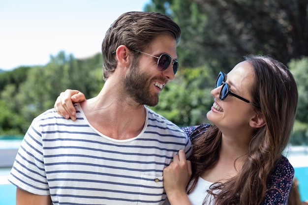 Feliz pareja en gafas de sol mirándose cerca de la piscina