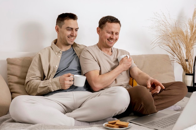 Feliz pareja gay con ropa informal pasando tiempo juntos en casa viendo películas en la computadora portátil y bebiendo té Dos hombres caucásicos relajando las relaciones homosexuales y el amor alternativo