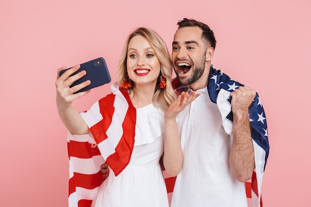 Feliz pareja hermosa que se encuentran aisladas sobre rosa, llevando la bandera americana, celebrando, tomando un selfie