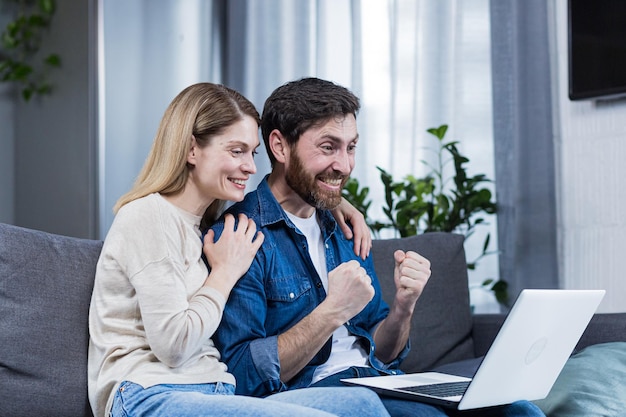 Feliz pareja hombre y mujer mirando la pantalla del portátil celebrando la victoria levantando las manos gesto de victoria y éxito