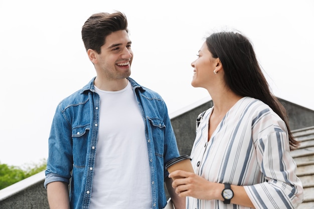 Feliz pareja hombre y mujer con vaso de papel sonriendo y hablando mientras baja las escaleras al aire libre