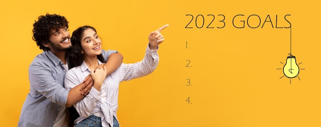 Foto feliz pareja india milenaria señala con el dedo la inscripción de objetivos 2023 con bombillas y lista de verificación