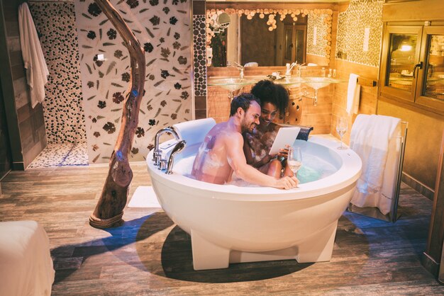 Feliz pareja interracial joven relajada disfrutando de sí mismos en el jacuzzi bebiendo vino y con una tableta