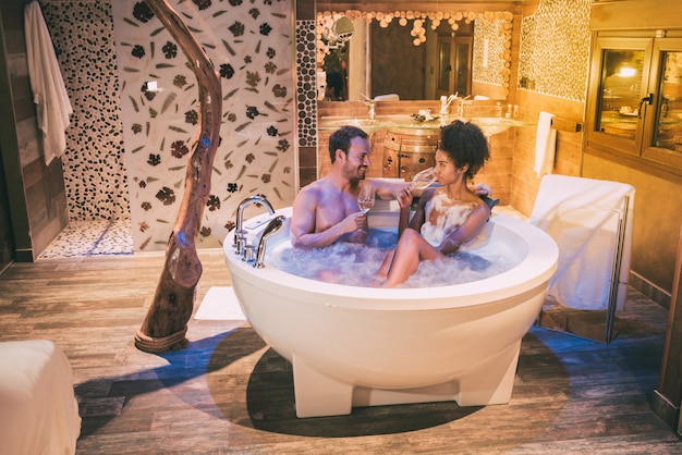 Feliz pareja interracial relajada en el jacuzzi disfrutando de su auto y bebiendo vino