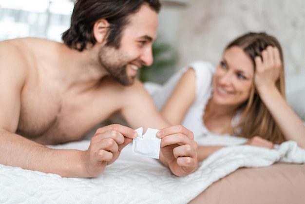 Feliz pareja joven acostada en la cama y abriendo el concepto de control de embarazo y ETS de enfoque selectivo del condón