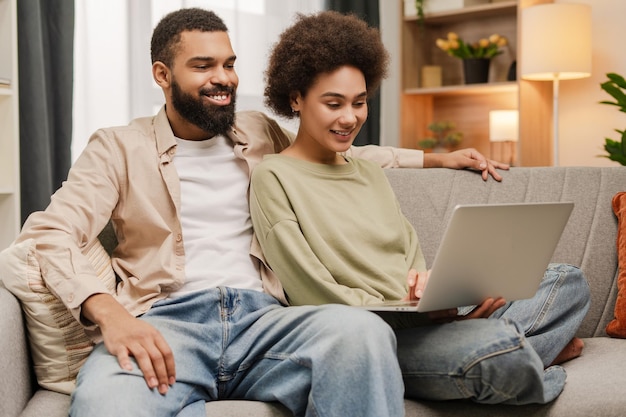 Una feliz pareja joven afroamericana sentada en el sofá de la sala de estar mientras usa una computadora portátil