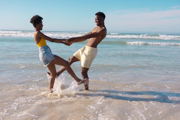Feliz pareja joven afroamericana tomándose de la mano y bailando en el mar contra el cielo en un día soleado