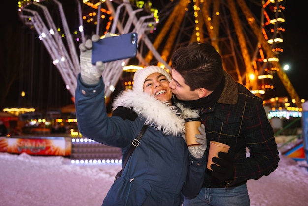 Feliz pareja joven alegre divirtiéndose en el parque de patinaje sobre hielo por la noche, sosteniendo tazas de café para llevar, tomando un selfie