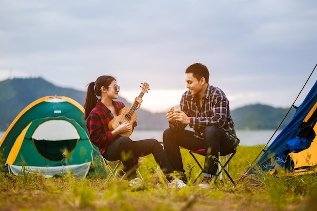 Feliz pareja joven asiática sentada en una silla de picnic bebiendo té y café mientras acampa junto al lago