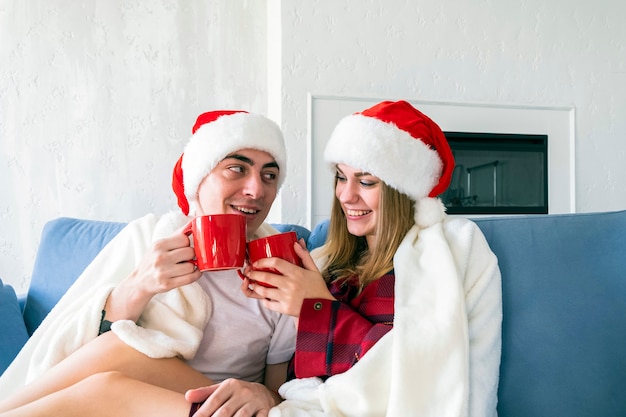 Feliz pareja joven beber té caliente o cacao sentado en casa en el sofá con gorros de santa
