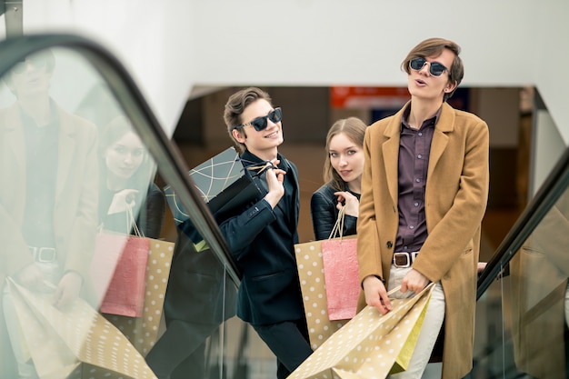 Feliz pareja joven con bolsas de compras bajando por las escaleras mecánicas y señalando con el dedo en el centro comercial