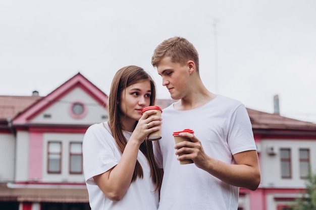 Feliz pareja joven camina por las calles de la ciudad y bebe café de una taza de cartón.