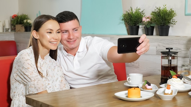 Feliz pareja joven chico chica hace selfie en smartphone moderno sentado en servido con tortas y bebidas en la mesa de café