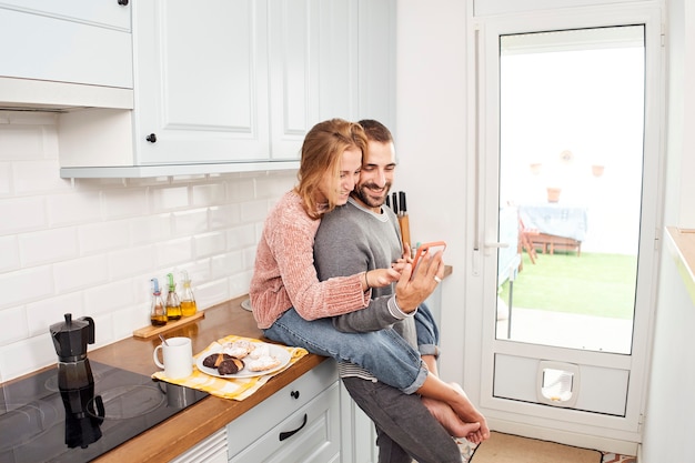 Feliz pareja joven en la cocina de casa, mediante teléfono móvil