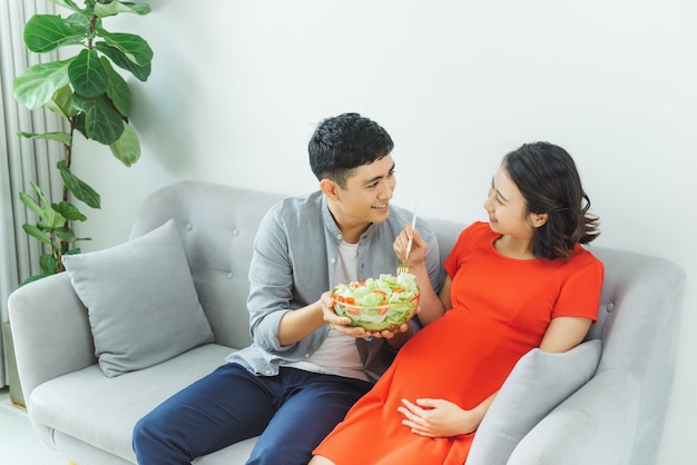 Feliz pareja joven comiendo ensalada juntos en el sofá en la sala de estar