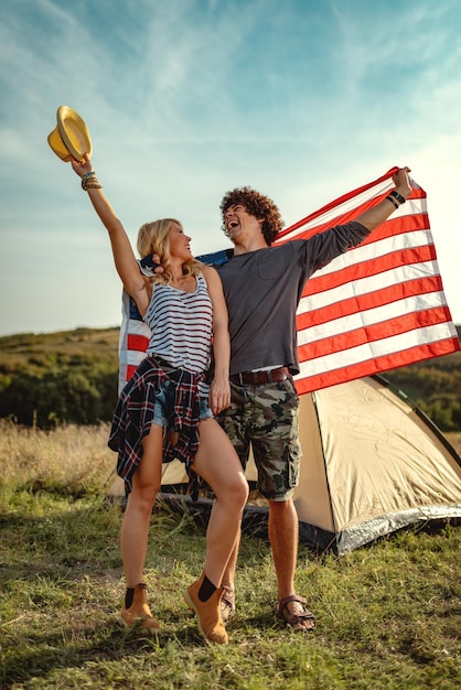 Feliz pareja joven disfruta de un día soleado en la naturaleza. Se están abrazando, sosteniendo una bandera estadounidense frente a una tienda de campaña.