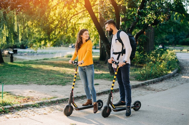 Feliz pareja joven disfrutando juntos mientras conduce patinetes eléctricos en el parque de la ciudad.