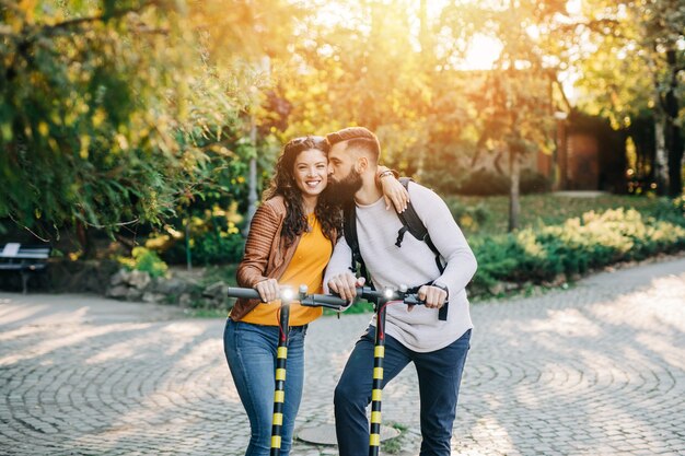 Feliz pareja joven disfrutando juntos mientras conduce patinetes eléctricos en el parque de la ciudad.