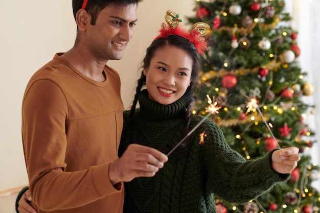 Feliz pareja joven diversa quemando luces de bengala en la celebración de Navidad en casa