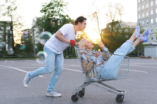 Foto feliz pareja joven divertirse con un carrito de la compra en el estacionamiento de un supermercado