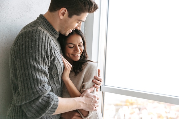 Feliz pareja joven encantadora de pie junto a la ventana en casa