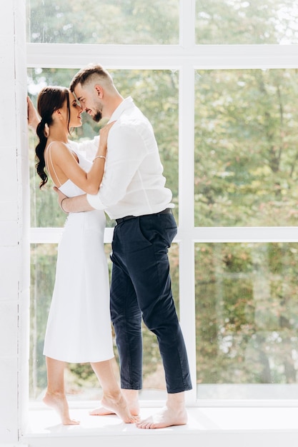 Feliz pareja joven encantadora de pie junto a la ventana en casa