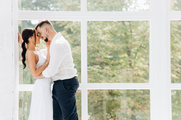 Feliz pareja joven encantadora de pie junto a la ventana en casa