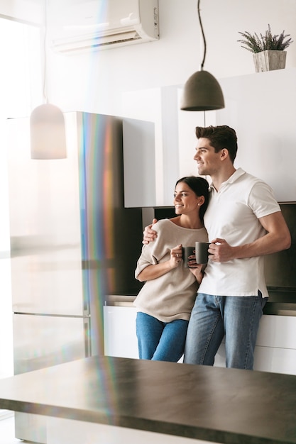 Foto feliz pareja joven encantadora sosteniendo tazas mientras está de pie en la cocina de casa