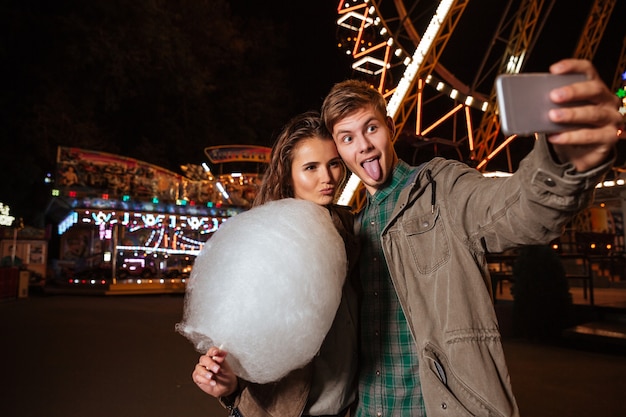 Feliz pareja joven haciendo muecas y tomando selfie en el parque de atracciones