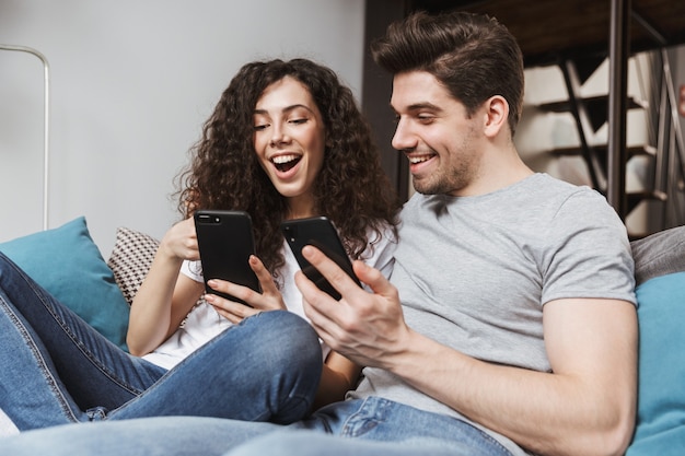 Feliz pareja joven hombre y mujer sentados en el sofá en casa y usando el teléfono inteligente juntos