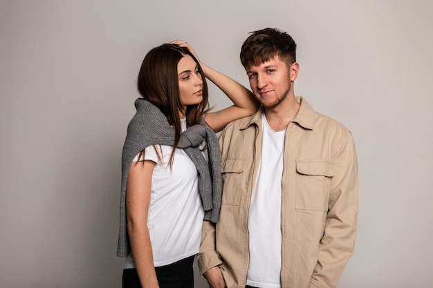 Feliz pareja joven de moda con elegante ropa exterior con camisetas blancas, una chaqueta beige y un suéter gris vintage en el estudio sobre un fondo gris. Look y estilo casual de ropa masculina y femenina.