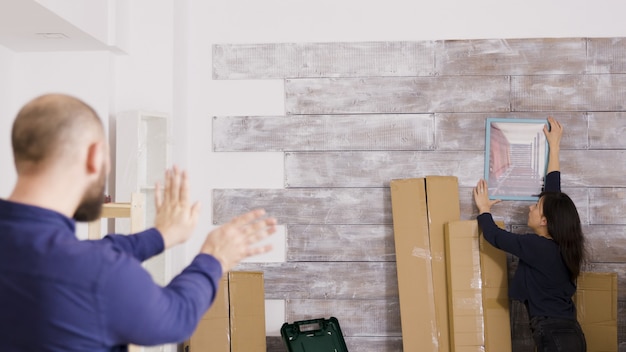 Feliz pareja joven mudarse a un nuevo apartamento colgando un cuadro en la pared. Trabajo en equipo de pareja.