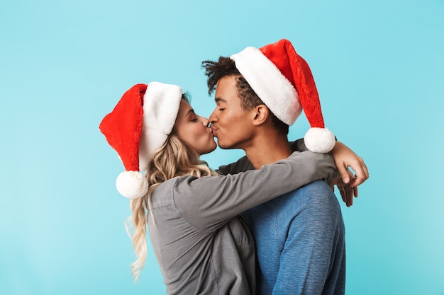 Feliz pareja joven multirracial con sombrero rojo de navidad aislado sobre pared azul, besos