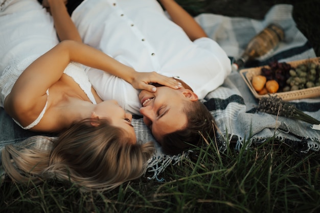 Feliz pareja joven en el parque se relaja en el picnic de verano. Ellos acostados en una manta sobre la hierba verde, mirándose y sonriendo.