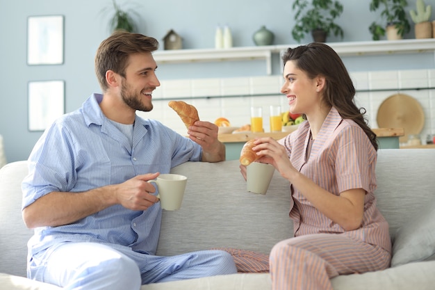 Feliz pareja joven en pijama en la cocina desayunando, alimentándose con un croissant.