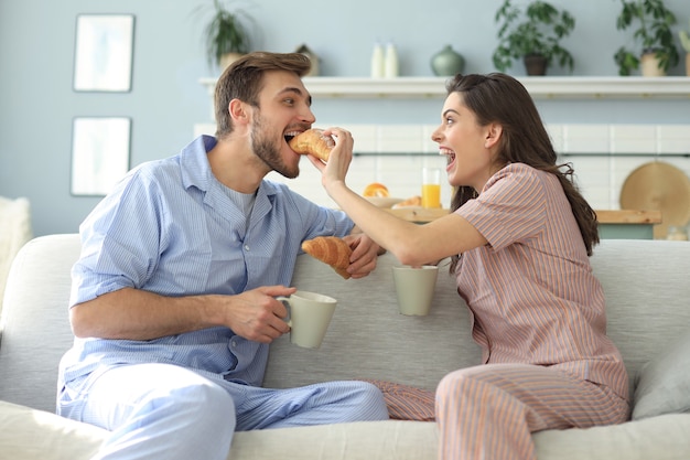 Feliz pareja joven en pijama en la cocina desayunando, alimentándose con un croissant.