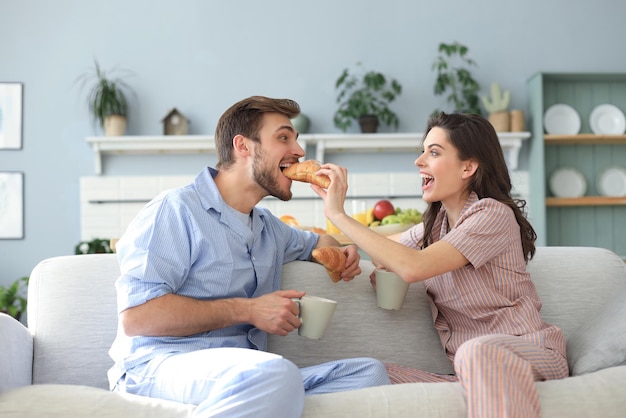 Feliz pareja joven en pijama en la cocina desayunando, dándose de comer un croissant.