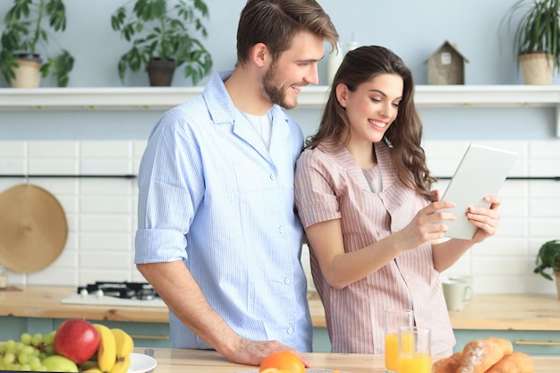 Feliz pareja joven en pijama viendo contenido en línea en una tableta didital y sonriendo en la cocina de casa.