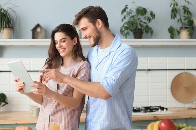 Feliz pareja joven en pijama viendo contenido en línea en una tableta didital y sonriendo en la cocina en casa.