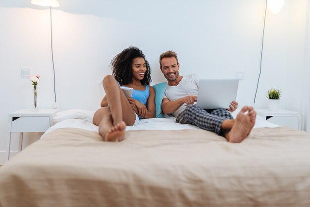 Foto feliz pareja joven relajado en casa acostado en la cama en la tableta y la computadora