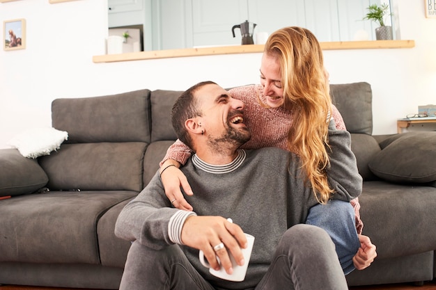 Feliz pareja joven relajándose hablando sosteniendo tazas de café en el sofá juntos en la sala de estar