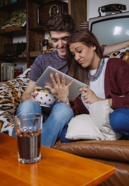 Feliz pareja joven riendo mientras mira tableta digital sentado en un sofá en casa