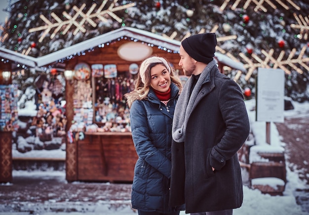 Feliz pareja joven con ropa abrigada parada cerca de un árbol de Navidad de la ciudad, disfrutando pasar tiempo juntos. Vacaciones, Navidad, Invierno.