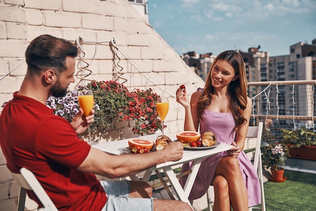 Feliz pareja joven en ropa casual desayunando y sonriendo mientras está sentado en el patio de la azotea al aire libre