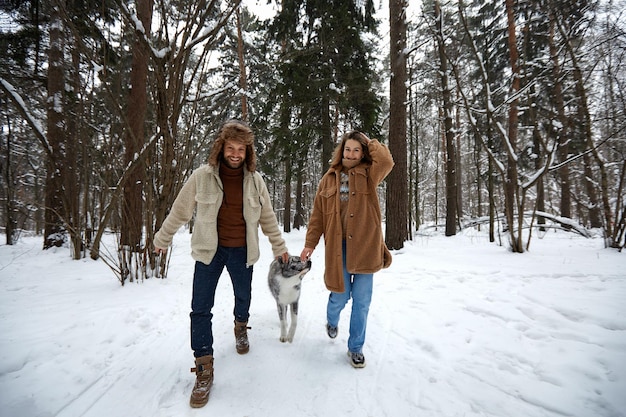 Feliz pareja joven en ropa de invierno jugando con husky siberiano de pura raza y divirtiéndose en un bosque de pinos nevados