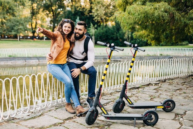 Feliz pareja joven con scooters eléctricos disfrutando juntos en el parque de la ciudad.