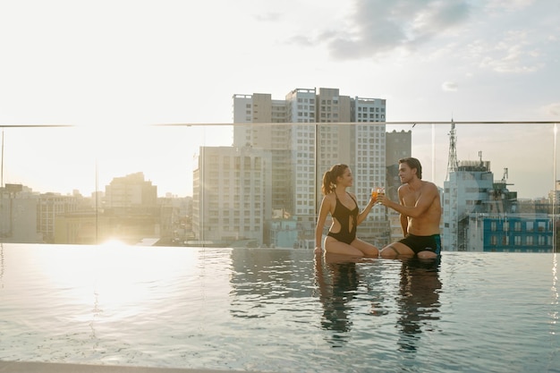 Feliz pareja joven sentada en el borde de la piscina y brindando con copas de cóctel