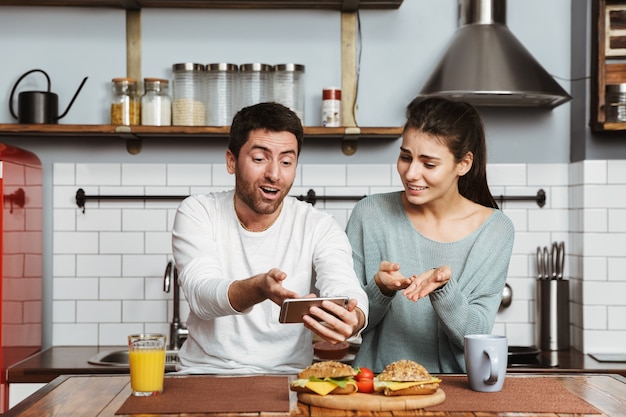 Feliz pareja joven sentada en la cocina durante el desayuno en casa, mediante teléfono móvil